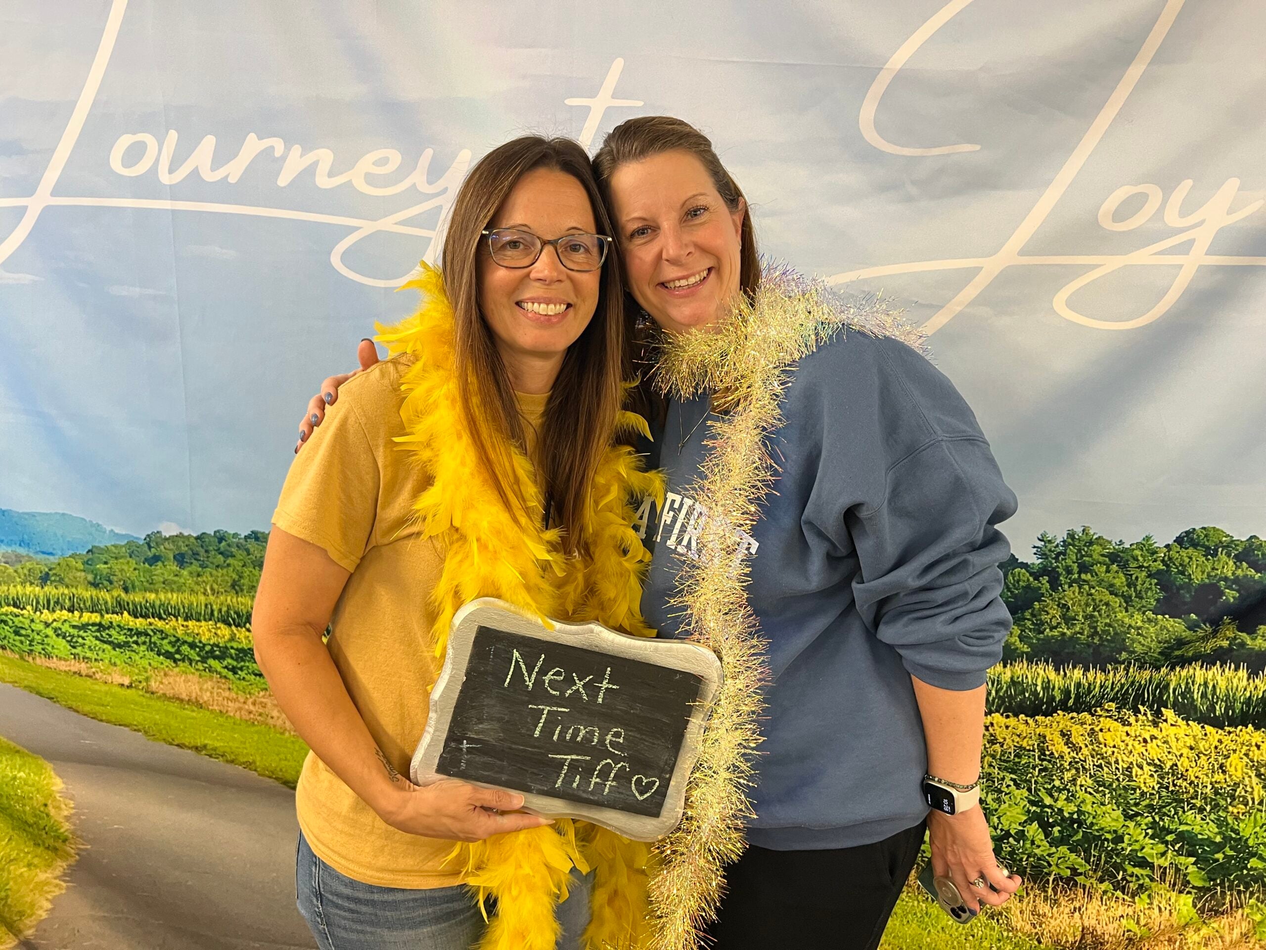 2 women smiling and huddling together with a yellow feathered boa