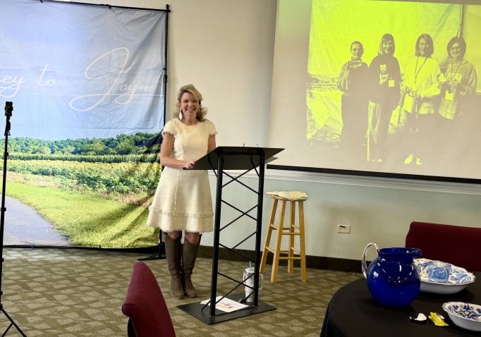 Lady stands at the front of a room in front of a projector. She's wearing a pink dress, black tights, and is smiling. She has blonde hair. She is teaching a group of women.