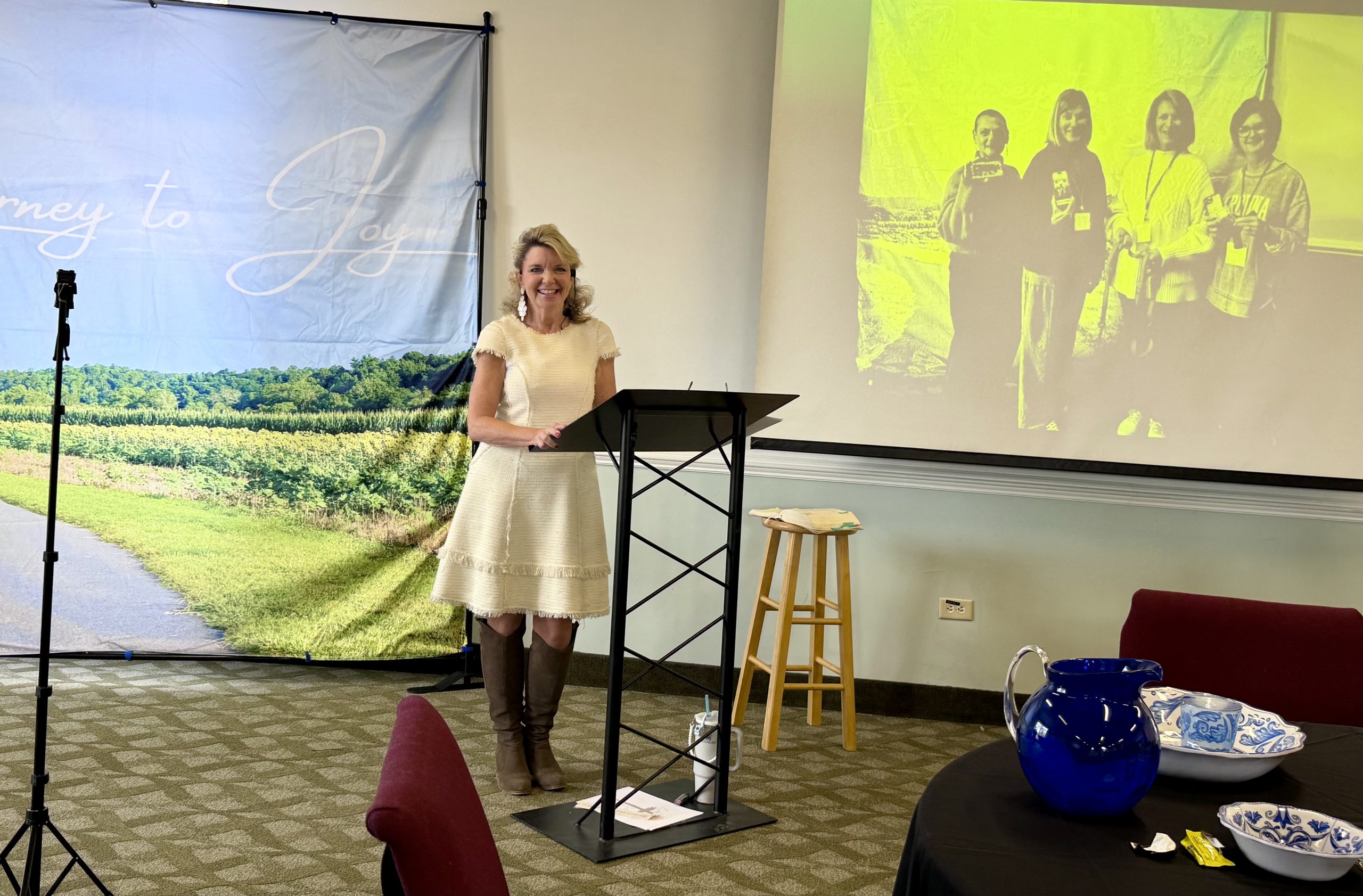 Lady stands at the front of a room in front of a projector. She's wearing a pink dress, black tights, and is smiling. She has blonde hair. She is teaching a group of women.